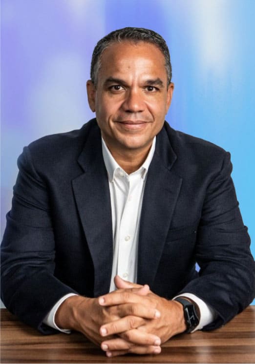 A man in a dark suit and white shirt sits at a wooden table with his hands clasped, smiling slightly at the camera against a blue and purple gradient background.