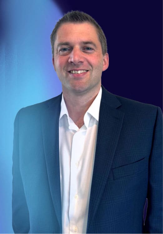 A man wearing a dark suit jacket and white dress shirt is smiling at the camera. The background is a gradient of deep blue and light blue tones.