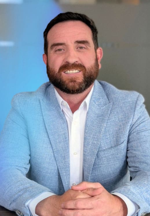 A man with short dark hair and a beard, wearing a light blue textured blazer and white shirt, sits at a table with his hands folded, smiling at the camera against a blurred background.